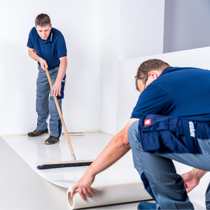 Zwei Männer in Arbeitsuniformen verlegen einen weißen Bodenbelag; der eine glättet das Material mit den Händen, während der andere es mit einem Besen glatt fegt.