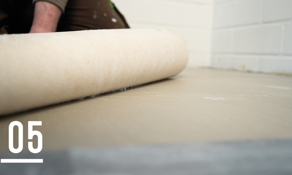 A close-up of a beige roll of material being unrolled onto a flat surface, with part of a person's arm visible in the background. The scene appears to be in a construction or renovation setting, with a white brick wall in the background. The number "05" is displayed in the bottom left corner.
