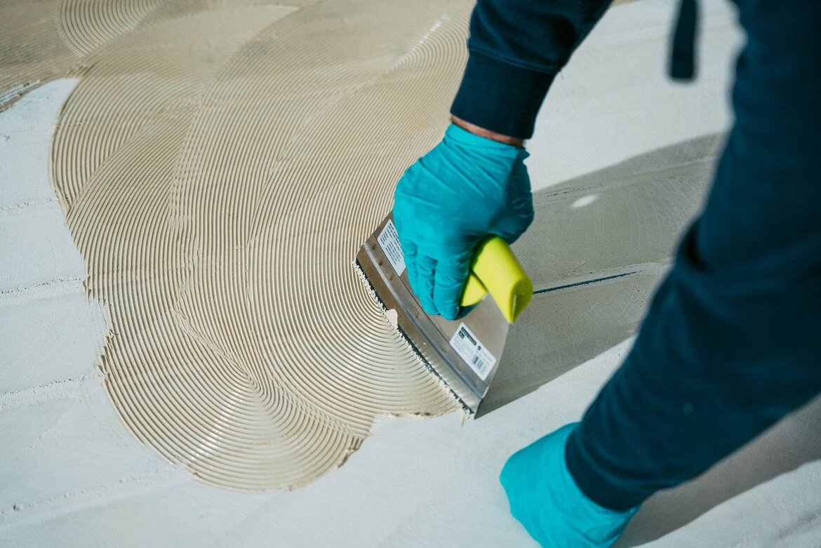A person wearing teal gloves spreads adhesive or mortar on a floor using a notched trowel, creating textured lines in the material.