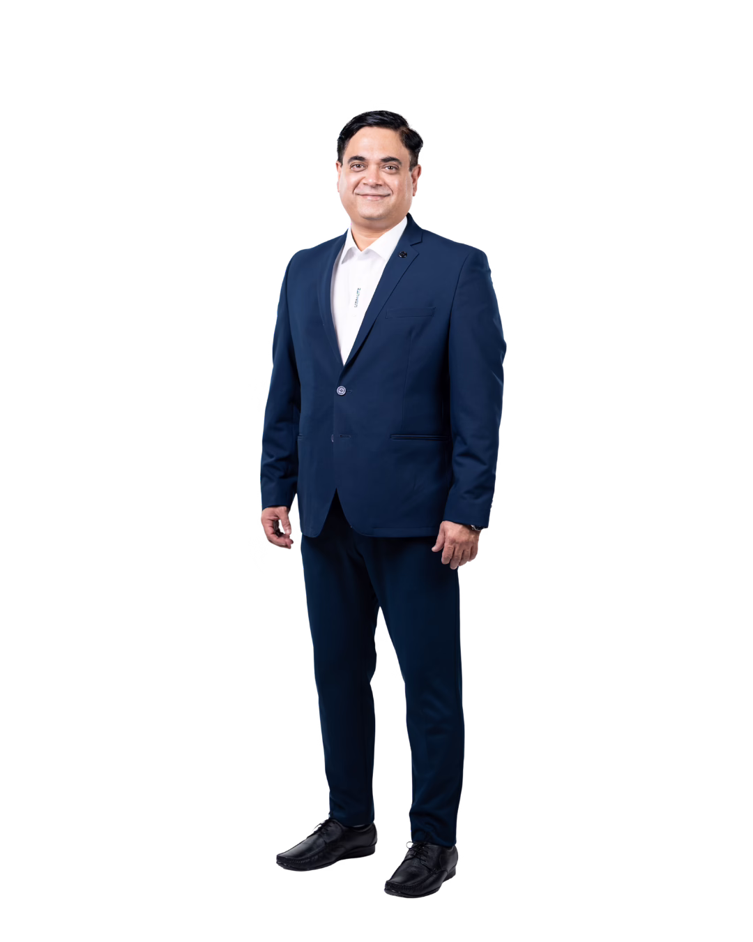 A man stands smiling, dressed in a navy blue suit, white collared shirt, and black shoes, against a plain white background.